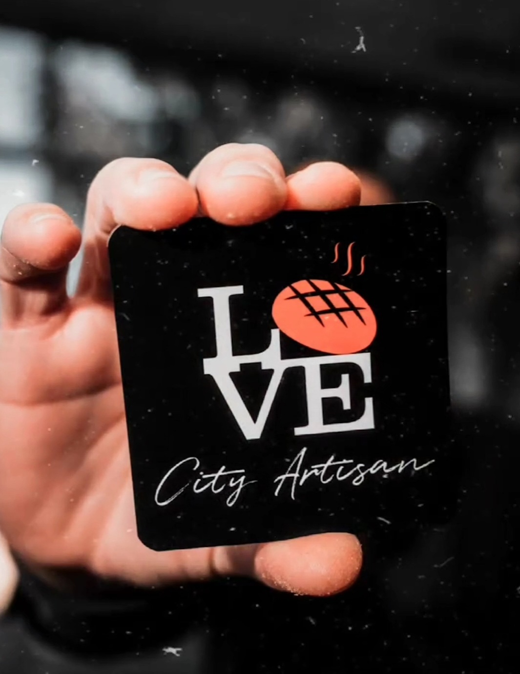 Hand holding a Love City Artisan logo card with a bread icon